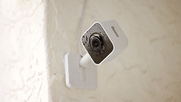 Rain drops on a Roku Outdoor Wired Camera mounted to the outside of a smart home